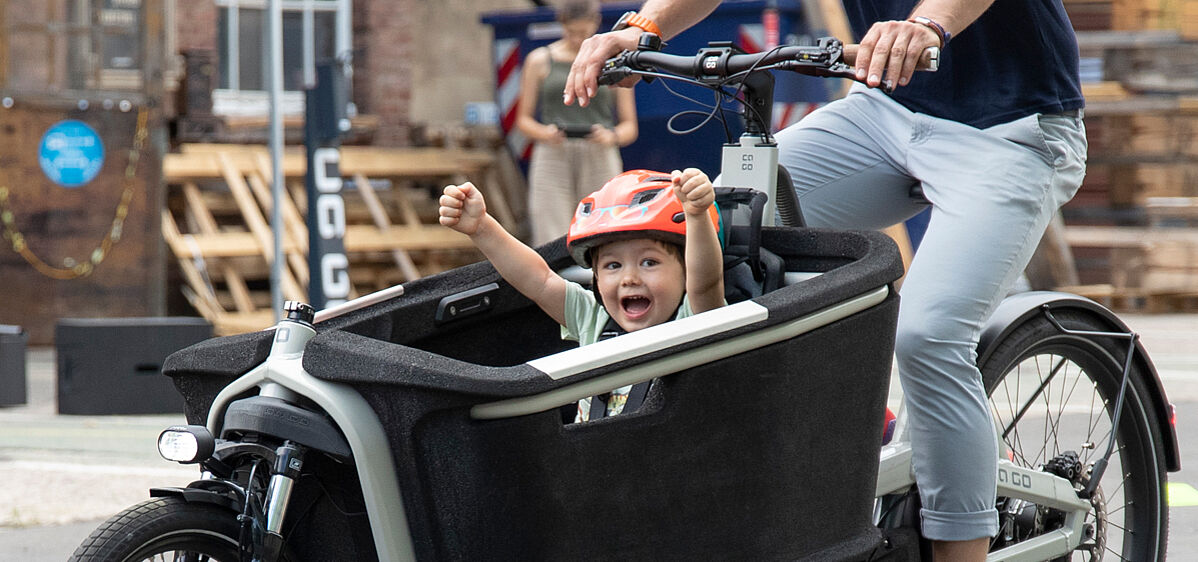 Ein Kind in einem Lastenrad hebt jubelnd die Arme, das Lastenrad fährt eine erwachsene Person in Jeans. für den Kindertransport ist der Traum vieler junger Familien.