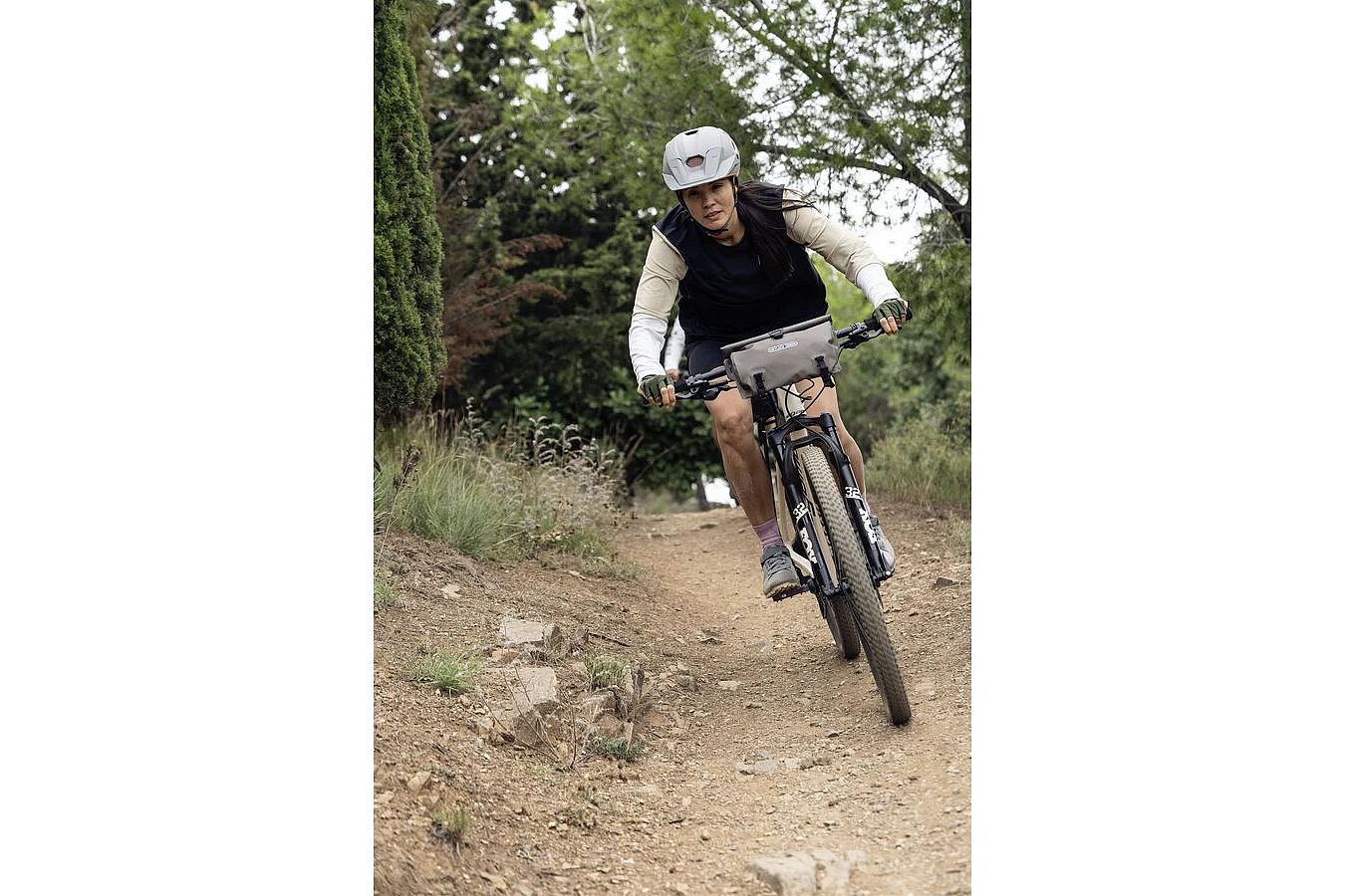 Person fährt mit einem Mountainbike über einen schmalen, steinigen Waldweg, eine Lenkertasche am Lenker. 
