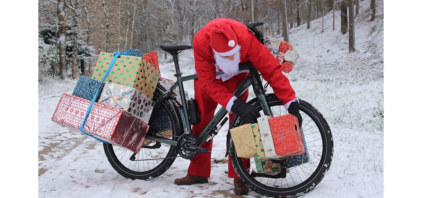 Man in Weihnachtskleidung beugt sich über ein mit Geschenken beladenes Fahrrad im Schnee. 
