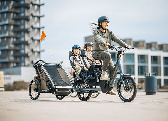 Frau fährt ein Lastenrad Longtail, auf dem langen Gepäckträger hinten sitzen zwei Kinder in je einem Kindersitz. Das Fahrrad zieht zudem einen Kinderfahrradanhänger.